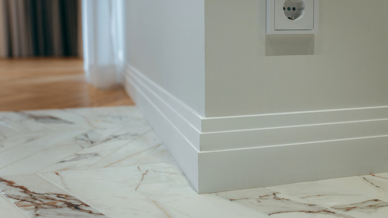 White baseboard molding with several ridges wraps around the corner of a wall against marble tile flooring.